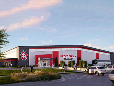 This photo shows an exterior artist rendering of the future Gassen Family Fieldhouse, courtesy of Bill and Jill Gassen and T. Denny Sanford. The building's exterior is red, black and white, with a giant USD Coyote logo on the lefthand side. It shows people entering and leaving the facility, as well as cars in the parking lot.