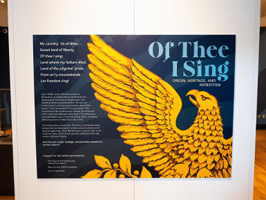 This photo shows a wall display in the National Music Museum introducing the temporary exhibit, "Of Thee I Sing." The exhibit description appears in white text against a blue background, while a large golden eagle with outstretched wings is prominently featured to the right.