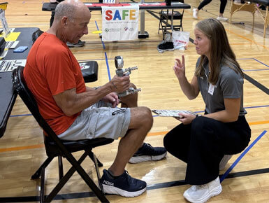 A woman crouches beside and speaks to a man, who is seated in a chair, as he squeezes a hand grip to test his strength during a fitness exam.