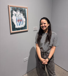 Mariana Astete stands next to a painting in an art gallery. She is smiling at the camera.