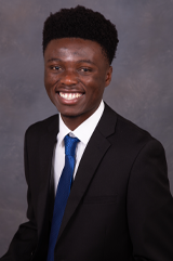 A headshot of Finiasi Epapy. He wears a black suit, white shirt and blue tie. He is sitting in front of a gray background. 