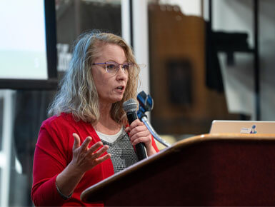 Angela Jackson stands at a podium, speaking into a microphone, and presenting on Digital Accessibility. She is wearing glasses and a red cardigan over a grey patterned dress,. She is gesturing with her right hand while looking toward an audience off-camera.