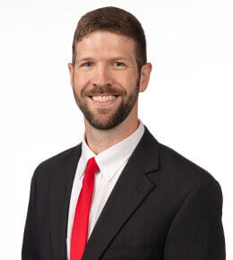 Jed Droge poses for his professional photo. He is wearing a black suit with a white undershirt and bright red tie.