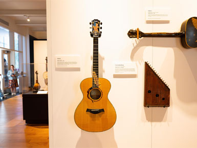 The Taylor Liberty Guitar sits mounted on a white wall in the National Music Museum. It is oak-ish brown, with the neck of the guitar displaying an etched, rolled-up document on it. The sound hole is bordered with a U.S. flag.