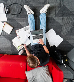 A USD student sits on the floor in a lounge, leaning against a red couch. He studying for classes, with his laptop on his lap and notes and books surrounding him.