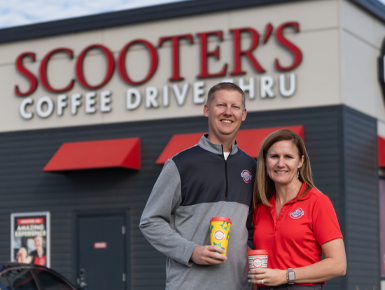Travis and Kristen Rhoades stand next to each other and hold coffee cups outside a Scooter's Coffee Drive Thru.