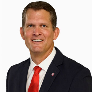 David Herbster headshot. He wears a black suit and a red tie.
