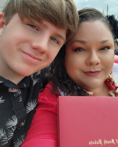 A woman poses with her son for a selfie. She is wearing graduation regalia and holding her diploma.