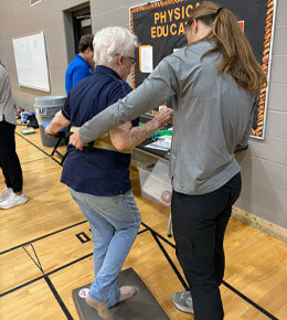 A physical therapist or trainer assists an older woman who is balancing on one foot on a padded mat during a movement evaluation.