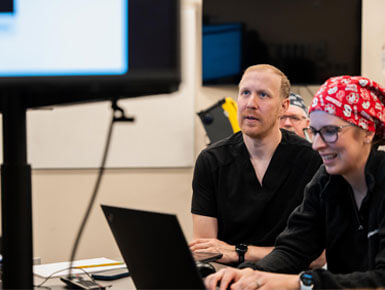 Two clinicians in black scrubs work at a laptop in a clinical setting; a woman in a red patterned cap smiles as her colleague watches attentively.