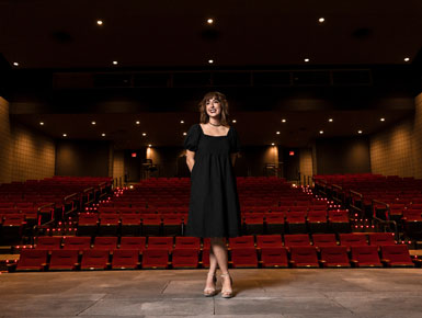 A wide shot portrait of Kylie Groves standing on stage in a theater. 