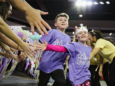 Two children wearing purple shirts run through a tunnel of people holding out their hands. The two children reach out their hands for high fives and smile. 
