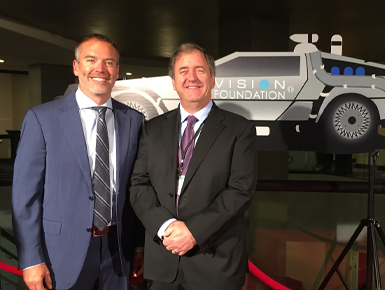 Two men in suits stand next to each other and smile in front of a gray car that says Vision Foundation on the side.