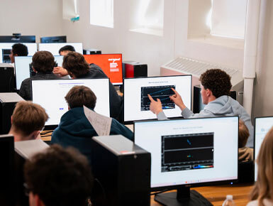 Several USD students sit in a classroom, with big computer monitors in front of them. Some students are pointing at their screens, while others are collaborating together.