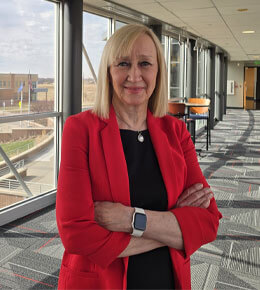 Cheryl Hartman stands in a hallway of a building on the USD - Sioux Falls campus. She is wearing a bright red blazer with a black shirt underneath.