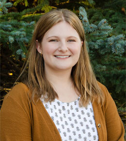 Anna Van Kley stands in front of a green evergreen tree, wearing a yellow cardigan and a white shirt that has a design on it. She is smiling at the camera.