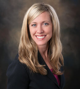 Erin Lehmann, Ed.D., poses for her headshot photo against a dark brown background. She is smiling at the camera and wearing a black blazer and pink shirt.