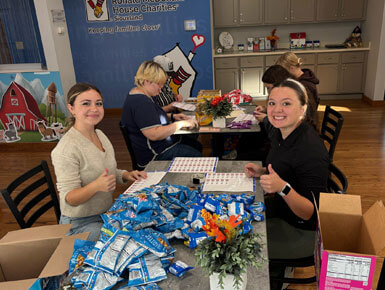 Two USD students sit at a table in a communal dining area, giving thumbs-up gestures to the camera. They are surrounded by a large pile of blue snack bags and sheets of labels, likely preparing care packages. A large "Ronald McDonald House Charities Siouxland" mural is visible on the wall in the background.