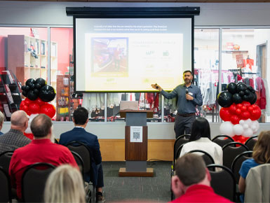 Anubhav Gupta, Ph.D., gives a presentation to an audience in the Muenster University Center. There are black, white and red balloons on either side of him, as well as a screen behind him and a podium to his right.