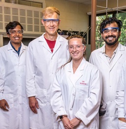 Group photo of Faculty Member and Students in White Lab Coats and Safety Glasses.