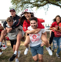 Students smiling at a tailgate and carrying each other around.